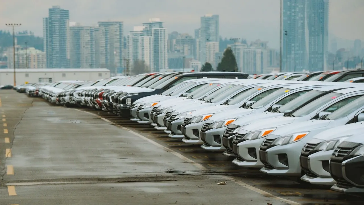 Rows of cars lined up for sale in an outdoor lot with a city skyline behind them