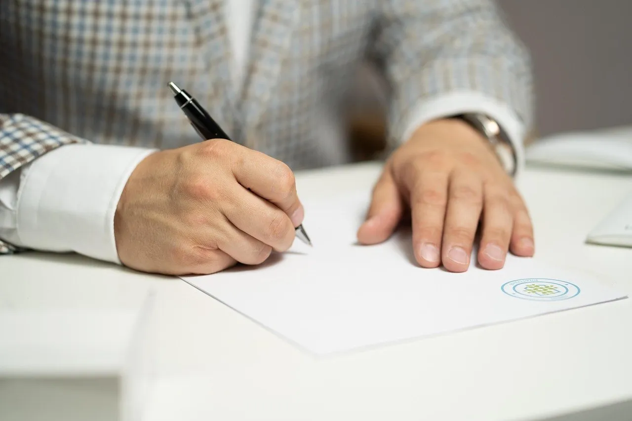 Person in a checked jacket signing a document with a pen at a white desk